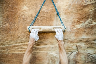 Two hands covered in chalk grip a wooden hangboard attached to a rope against a textured rock wall.