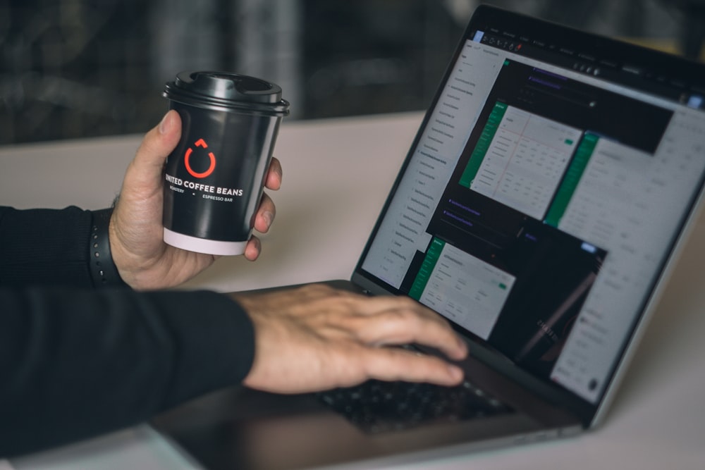 a person holding a cup of coffee in front of a laptop