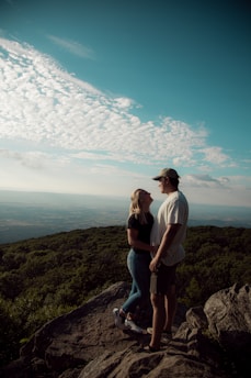 A couple standing on a scenic mountain overlook, gazing at a vast landscape of rolling hills and valleys.