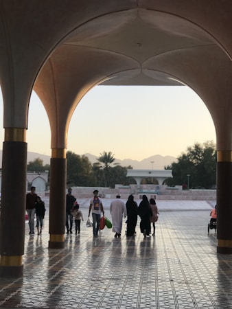 A group of people walking under a large, archway with intricate floor tiles reflecting the light. In the background, there are trees and distant mountains under a soft, evening sky.