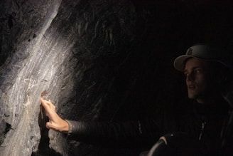A person wearing a dark jacket and a cap is touching or inspecting a textured, illuminated rock surface in a dimly lit environment. The focused light creates dramatic shadows and highlights on the rock face.