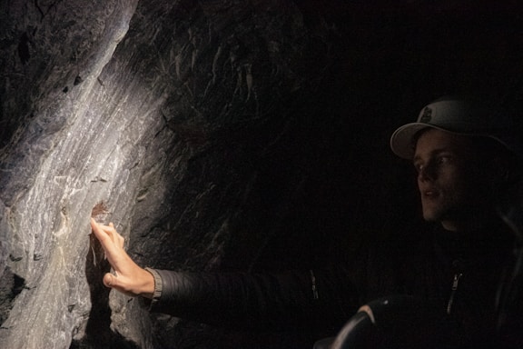 A person wearing a dark jacket and a cap is touching or inspecting a textured, illuminated rock surface in a dimly lit environment. The focused light creates dramatic shadows and highlights on the rock face.