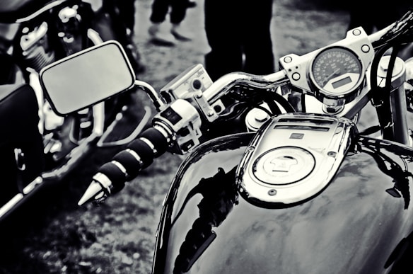 A close-up view of a motorcycle's dashboard, featuring the speedometer, fuel gauge, and handlebars. The image appears in black and white, emphasizing the chrome details and textures of the motorcycle, with a side mirror reflecting part of the environment.