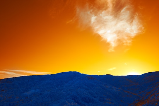 A vibrant landscape showcasing a mountain range in blue hues beneath a striking orange sky. The clouds are illuminated with shades of white and wispy patterns, contrasting sharply with the vivid warm tones of the sunset.