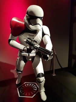 A life-sized model of a futuristic soldier in a white and black armored suit, posed in a dynamic stance holding a large weapon. The helmet is sleek and featureless, with a red shoulder pad accentuating its stark design. The figure is positioned against a dark backdrop with red lighting, adding a dramatic effect.