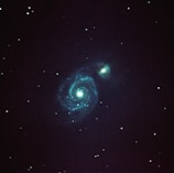 A spiral galaxy with well-defined arms is prominently visible against a dark starry background. Bright turquoise and blue hues illuminate the central core and spiral patterns, surrounded by numerous small stars scattered throughout the blackness of space.
