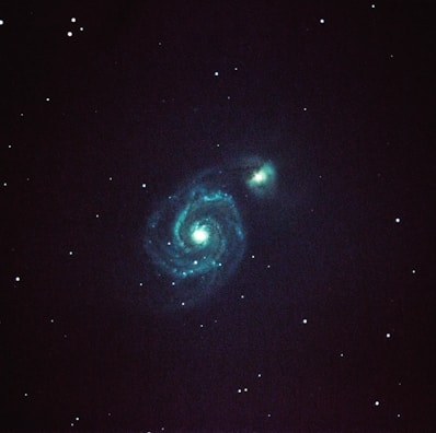 A spiral galaxy with well-defined arms is prominently visible against a dark starry background. Bright turquoise and blue hues illuminate the central core and spiral patterns, surrounded by numerous small stars scattered throughout the blackness of space.