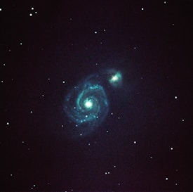 A spiral galaxy with well-defined arms is prominently visible against a dark starry background. Bright turquoise and blue hues illuminate the central core and spiral patterns, surrounded by numerous small stars scattered throughout the blackness of space.