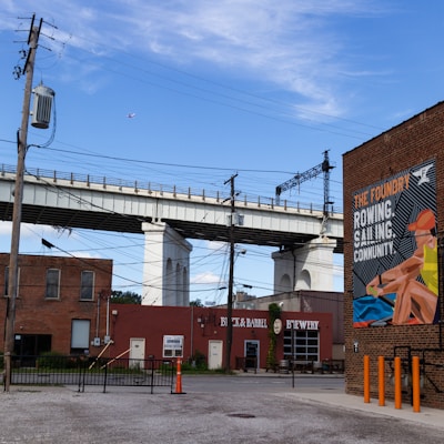 An urban scene featuring a large bridge structure elevated over a street. Below the bridge are several brick buildings, including a brewery and a colorful mural depicting a rowing scene with bold text promoting community activities. Electrical wires crisscross the area, and there are several utility poles. A plane is visible in the sky, adding a sense of movement.