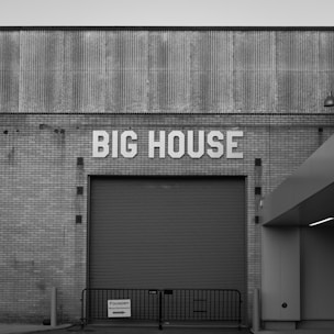 A large roller door is set into a brick building facade with the words 'BIG HOUSE' displayed prominently above. The architecture features a combination of corrugated and brick textures, creating a vintage industrial aesthetic. A small sign reading 'FOUNDRY Main Entrance' is affixed to a temporary railing in front.