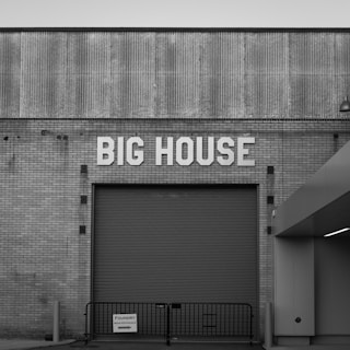 A large roller door is set into a brick building facade with the words 'BIG HOUSE' displayed prominently above. The architecture features a combination of corrugated and brick textures, creating a vintage industrial aesthetic. A small sign reading 'FOUNDRY Main Entrance' is affixed to a temporary railing in front.