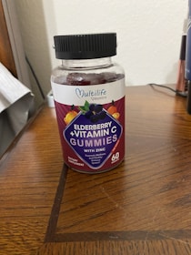 A plastic bottle of Multilife Vitamins featuring elderberry and vitamin C gummies with zinc is placed on a wooden surface. The label prominently displays images of elderberries and citrus fruits with the text indicating 60 gummies per bottle. The overall setting appears to be indoors, likely on a table or desk.