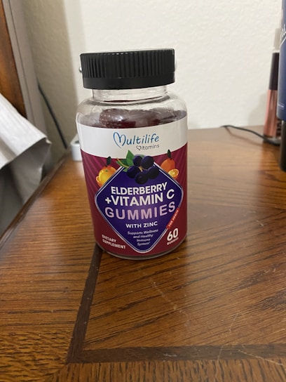 A plastic bottle of Multilife Vitamins featuring elderberry and vitamin C gummies with zinc is placed on a wooden surface. The label prominently displays images of elderberries and citrus fruits with the text indicating 60 gummies per bottle. The overall setting appears to be indoors, likely on a table or desk.