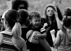 A black and white image of a group of people, with a focal point on a crying child being held by an adult. The child appears to be in distress, while the surrounding adults and children have various expressions, some looking away or engaged in other activities.