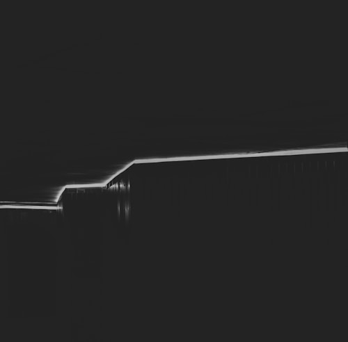 A minimalistic black and white photograph featuring a diagonal line created by lighting or shadow, dividing the image. The simplicity of the composition emphasizes geometric shapes and abstract forms.