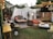 An outdoor seating area styled with a bohemian vibe, featuring an assortment of comfortable couches, colorful cushions, and various furniture pieces such as tables and a wooden barrel. The space is adorned with string lights overhead, white fabric drapes, and greenery, creating a cozy and inviting atmosphere. A chandelier hangs for added elegance, while a green lawn provides the base for the eclectic decor.