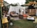 An outdoor seating area styled with a bohemian vibe, featuring an assortment of comfortable couches, colorful cushions, and various furniture pieces such as tables and a wooden barrel. The space is adorned with string lights overhead, white fabric drapes, and greenery, creating a cozy and inviting atmosphere. A chandelier hangs for added elegance, while a green lawn provides the base for the eclectic decor.