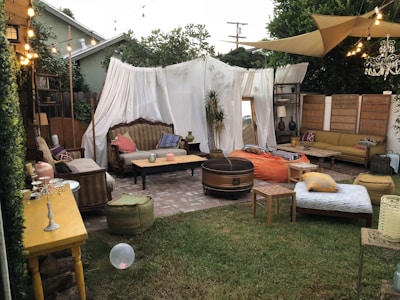 An outdoor seating area styled with a bohemian vibe, featuring an assortment of comfortable couches, colorful cushions, and various furniture pieces such as tables and a wooden barrel. The space is adorned with string lights overhead, white fabric drapes, and greenery, creating a cozy and inviting atmosphere. A chandelier hangs for added elegance, while a green lawn provides the base for the eclectic decor.