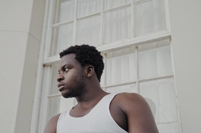 A person is gazing thoughtfully off to the side, standing in front of a large, multi-paned window. They are wearing a white tank top and appear to be in a calm, contemplative state.