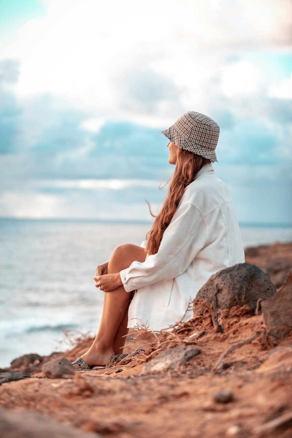 woman in white long sleeve shirt and white hat sitting on brown rock near body of on on on on