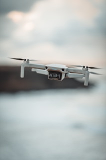 A drone flying in mid-air against a blurred, cloudy background. The drone appears to be hovering steadily, with its propellers in motion.