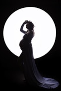 A silhouette of a pregnant woman standing gracefully in front of a large circular light source. She is wearing a long, flowing gown and a flower crown, with one hand resting on her belly and the other lifted to her head.