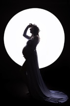 A silhouette of a pregnant woman standing gracefully in front of a large circular light source. She is wearing a long, flowing gown and a flower crown, with one hand resting on her belly and the other lifted to her head.
