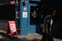 A storefront with a blue door and window frames. There are two wall-mounted lanterns flanking the door. Signs and posters, including one advertisement featuring a watermelon drink, are displayed on the walls near the entrance. A red A-frame signboard with the word 'MOISSON' is placed on the sidewalk. A person wearing a white and green shirt is walking past the store.