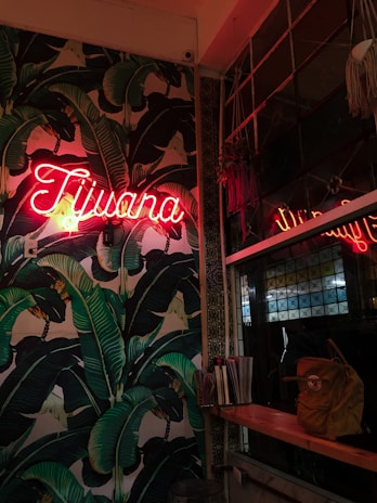 A vibrant interior wall adorned with a repeating pattern of large green banana leaves. A neon sign, casting a red glow, displays the word 'Tijuana'. To the right, a wooden shelf holds a backpack and some stacked booklets. A large window with a geometric design reflects part of the scene.