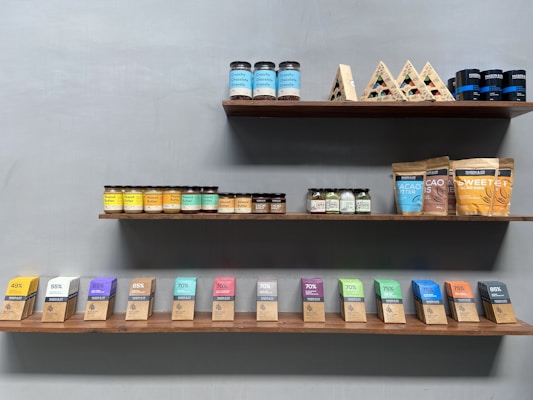 Shelves displaying a variety of packaged food products, including jars of peanut butter and almond butter, and assorted boxes and bags of cacao butter and sweeteners. The arrangement is neat and organized by product type and color, with a minimalist design.