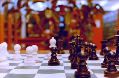 A close-up of a chessboard with chess pieces in focus, featuring both black and white pieces. The background appears blurred, suggesting a setting in a warmly lit room with soft, ambient light and indistinct decor.