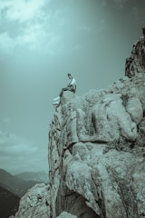 A person sits on the edge of a rocky cliff, looking out over a mountainous landscape. The scene conveys a sense of solitude and adventure, with the sky partly cloudy overhead.