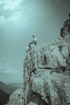 A person sits on the edge of a rocky cliff, looking out over a mountainous landscape. The scene conveys a sense of solitude and adventure, with the sky partly cloudy overhead.