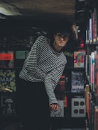 A person with curly hair and glasses is leaning slightly forward in a dimly lit room. They are wearing a black and white striped shirt. The background is filled with shelves of books that include titles related to Andy Warhol, Alfred Hitchcock, and Batman.