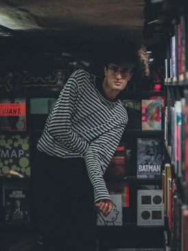 A person with curly hair and glasses is leaning slightly forward in a dimly lit room. They are wearing a black and white striped shirt. The background is filled with shelves of books that include titles related to Andy Warhol, Alfred Hitchcock, and Batman.