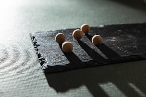 Five small spherical objects are placed on a rectangular piece of slate. The light creates elongated shadows, emphasizing the rough texture of the slate and the smooth surfaces of the spheres.