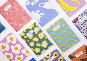 A collection of colorful notebooks arranged on a white surface. Each notebook features a unique and vibrant abstract design, including patterns of bananas, fried eggs, milk splashes, and other geometric shapes.