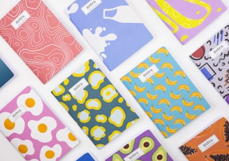 A collection of colorful notebooks arranged on a white surface. Each notebook features a unique and vibrant abstract design, including patterns of bananas, fried eggs, milk splashes, and other geometric shapes.