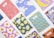 A collection of colorful notebooks arranged on a white surface. Each notebook features a unique and vibrant abstract design, including patterns of bananas, fried eggs, milk splashes, and other geometric shapes.