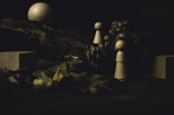 Wooden figurines are set among natural elements, such as moss and leaves, with a small group of mushrooms in the background. The scene is dimly lit, creating a mysterious and earthy atmosphere.