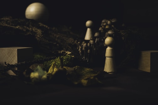 Wooden figurines are set among natural elements, such as moss and leaves, with a small group of mushrooms in the background. The scene is dimly lit, creating a mysterious and earthy atmosphere.