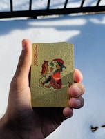 A person's hand holds a gold-colored playing card featuring an illustration of a joker with a bird perched on its hand. The card has a decorative, sparkling texture and the word 'Joker' printed in red. In the background, there is a snowy surface with shadows cast by a railing.