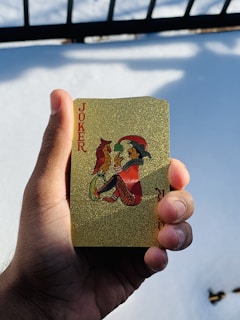 A person's hand holds a gold-colored playing card featuring an illustration of a joker with a bird perched on its hand. The card has a decorative, sparkling texture and the word 'Joker' printed in red. In the background, there is a snowy surface with shadows cast by a railing.