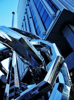 A modern architectural structure featuring a geometric metallic sculpture in the foreground. The background includes a contemporary building with glass windows reflecting the sky. The sculpture comprises interlocking shiny metal beams, creating a complex and abstract visual element.