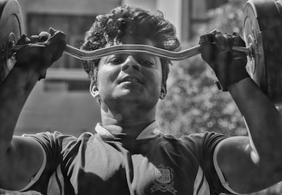A person is lifting a barbell close to their face, showing intense focus and strength. The individual is wearing gloves and a sports shirt and is outdoors, as indicated by the blurry background.