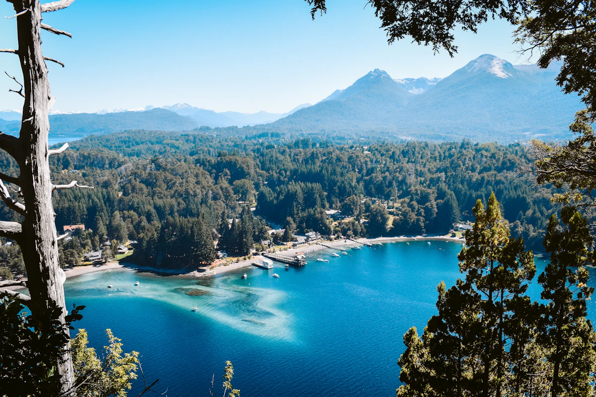 View through trees over the turquoise bay of Lake Nahuel Huapi at Villa La Angostura, with forested shores and mountains behind.