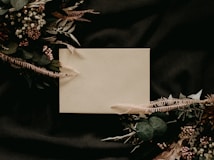 A plain, beige rectangular card is placed on a dark fabric background, surrounded by an arrangement of dried foliage and flowers. The foliage includes green leaves, white and pink small flowers, and beige fern-like elements, creating a rustic, natural frame around the card.