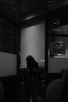 A person with a hunched posture is seated alone at a table in a dimly lit room. The scene is captured through a glass partition, giving the impression of distance and isolation. The room appears to be a public place like a café or waiting area, indicated by visible furniture and a no smoking sign. The image is in black and white, enhancing the mood of solitude and quiet reflection.