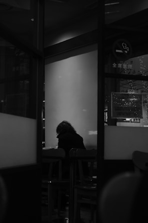 A person with a hunched posture is seated alone at a table in a dimly lit room. The scene is captured through a glass partition, giving the impression of distance and isolation. The room appears to be a public place like a café or waiting area, indicated by visible furniture and a no smoking sign. The image is in black and white, enhancing the mood of solitude and quiet reflection.