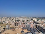 A panoramic view of a cityscape featuring numerous mid-rise apartment buildings under a clear blue sky. There is a construction site in the foreground with several unfinished structures and construction materials scattered around. Buildings in the background are densely packed, with a mix of residential and commercial properties.
